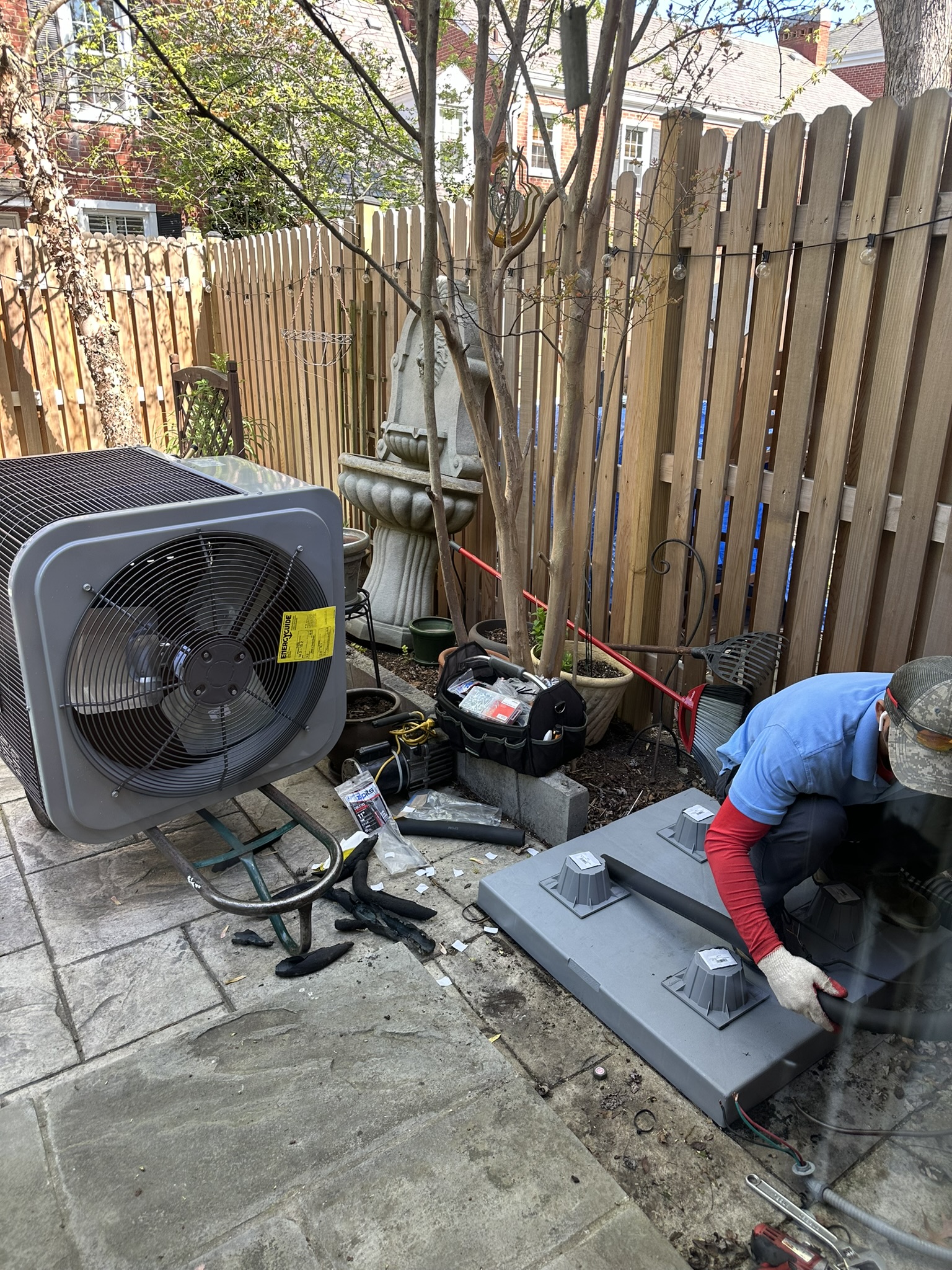 HVAC technician working on outdoor air conditioning equipment beside a wooden fence.